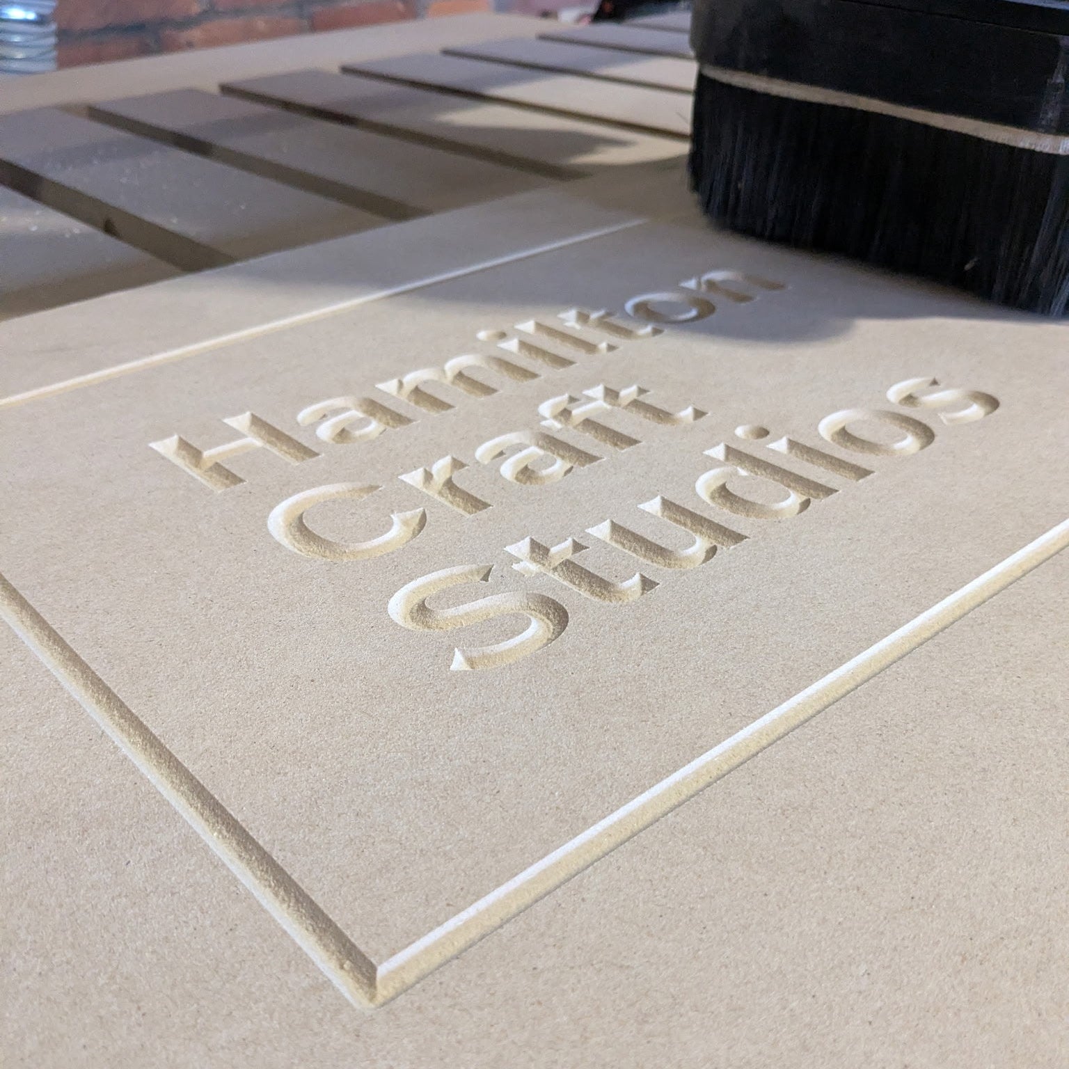 Beige surface (mdf) with 'Hamilton Craft Studios' engraved on it, with a blurred background.