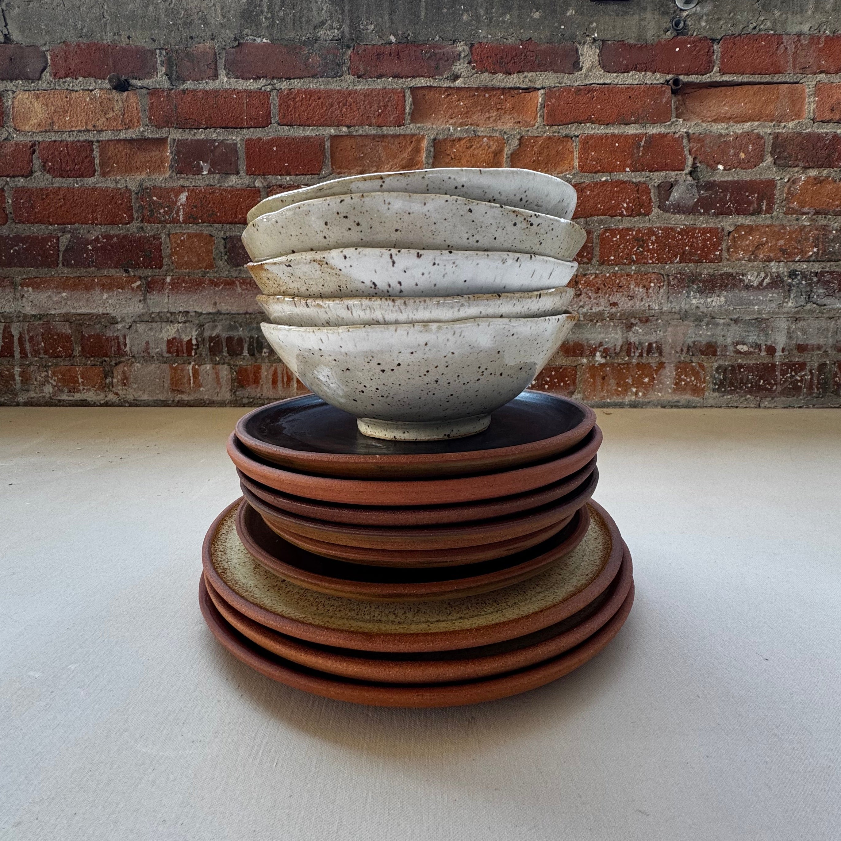A sculptural ceramic wall light fixture mounted on a white wall, featuring a textured off-white ribbed shade and a tiered base made of terracotta and brown ceramic rings, set against a brick column.