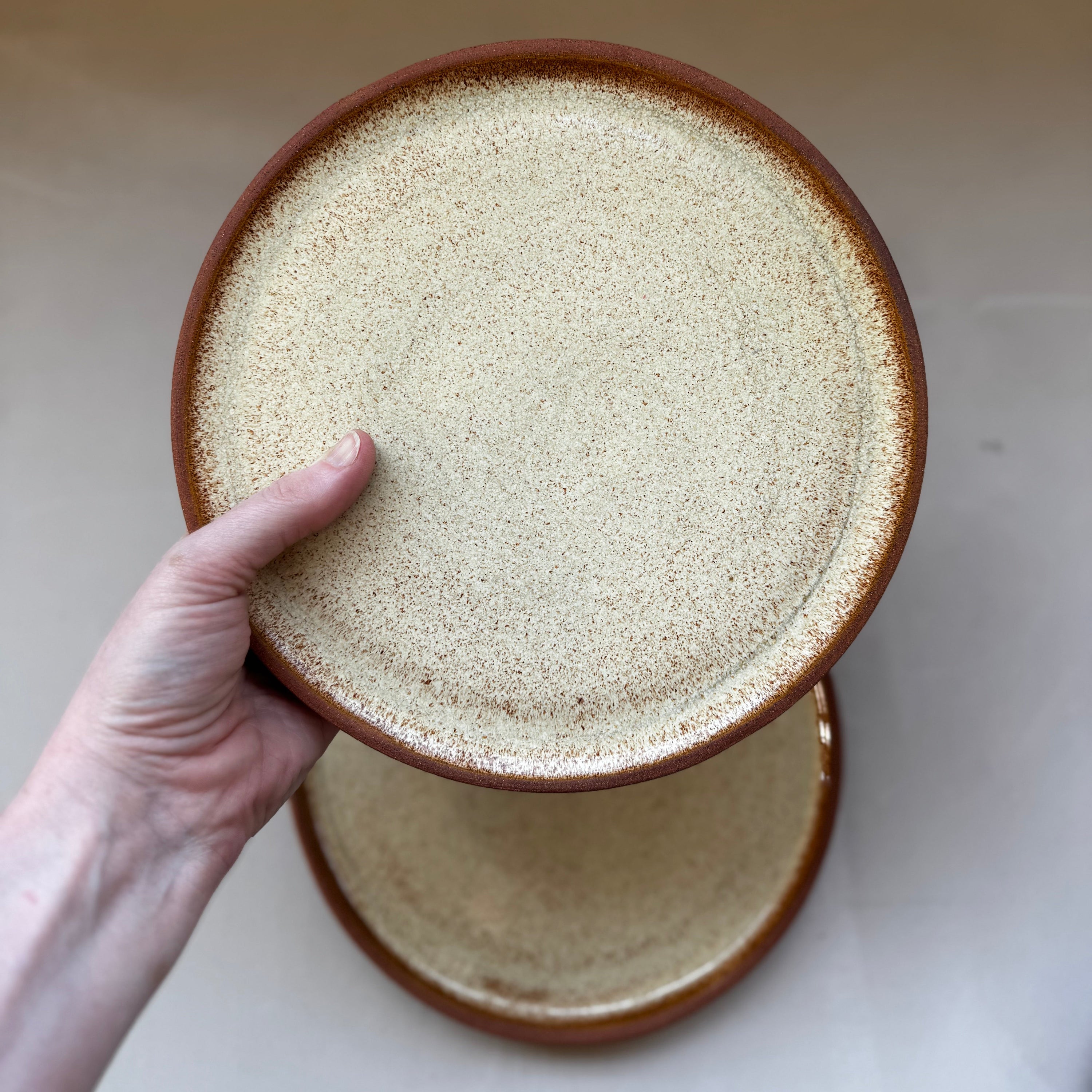 A hand holding a round ceramic plate with a speckled cream-beige glaze and a brown rim, with a second matching plate stacked below on a light surface.