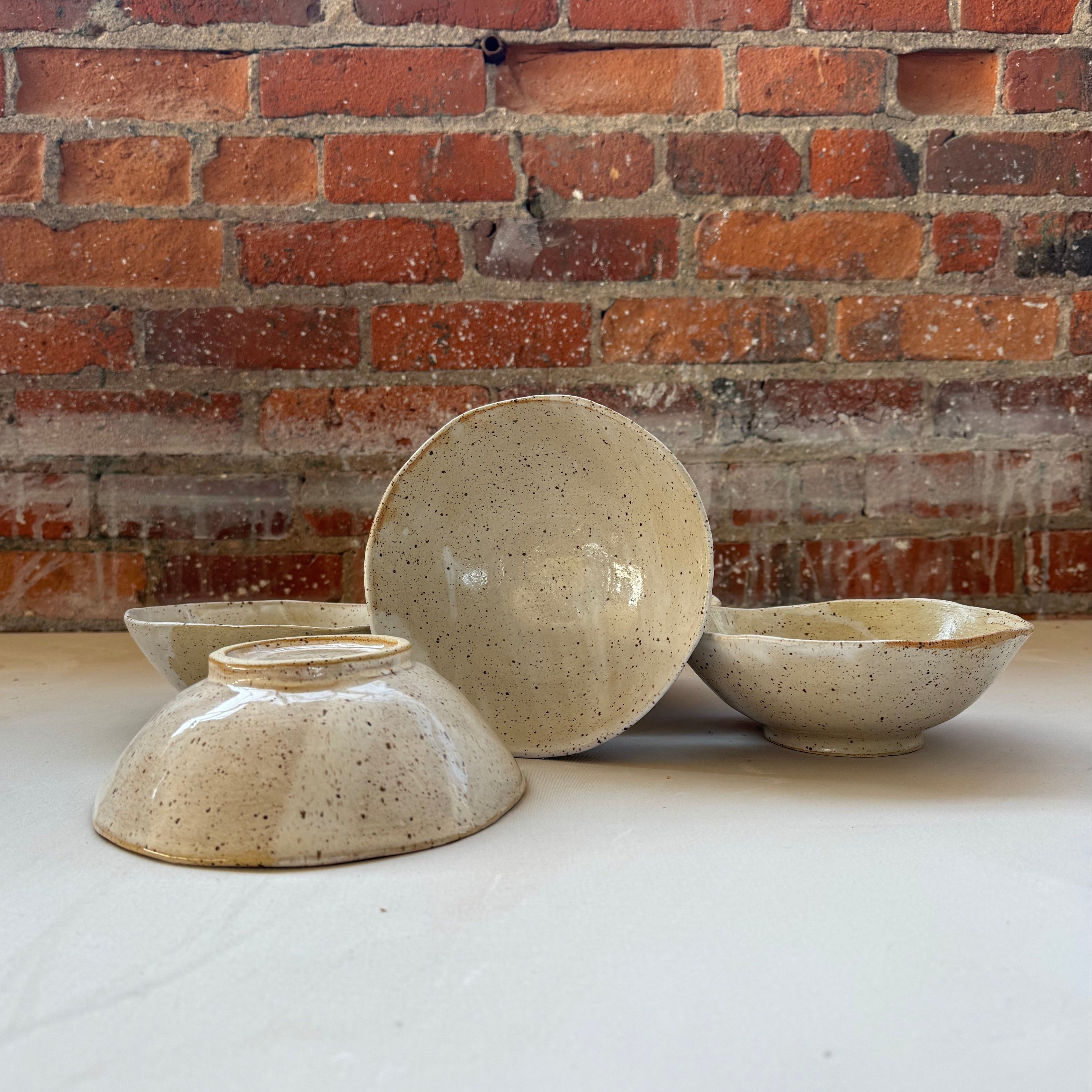 Four handmade ceramic bowls in a speckled beige glaze, arranged on a white surface with a rustic red brick wall background.