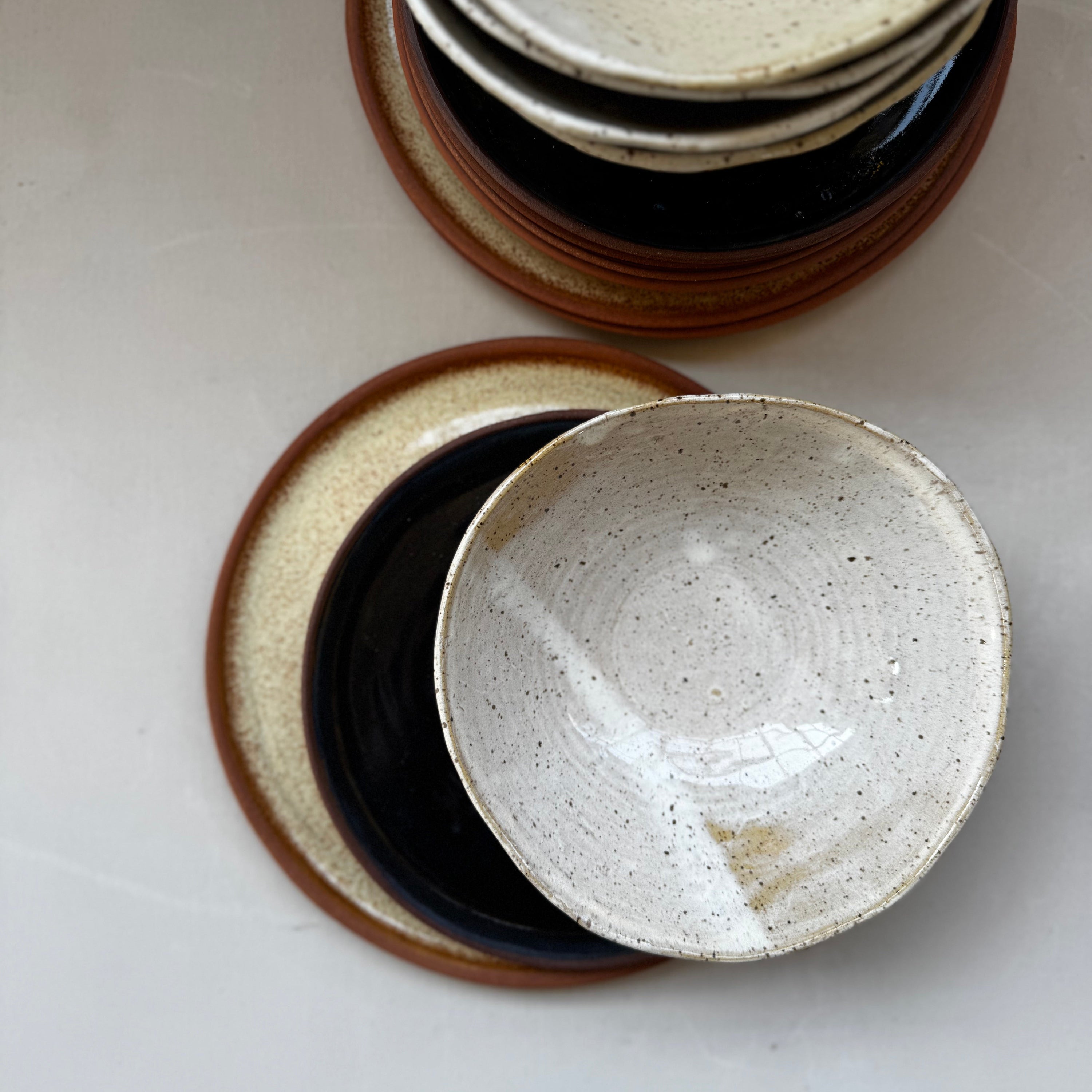 A stack of handmade ceramic bowls and plates in a speckled cream glaze with brown rims, arranged on a light neutral surface.