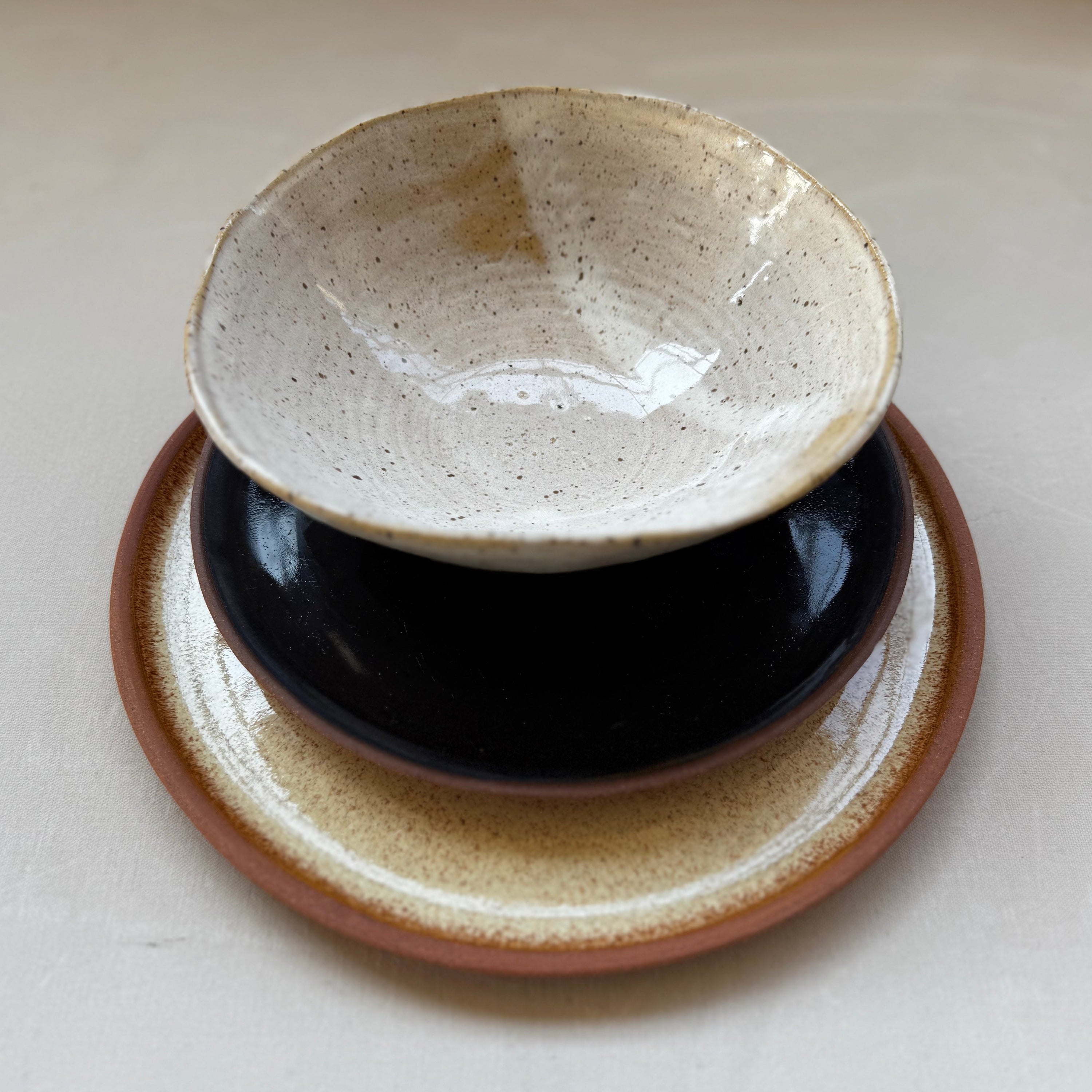 A handmade ceramic bowl with a speckled cream glaze sits on top of a glossy black saucer, which is placed on a matching brown and cream ceramic coaster, all set against a warm off-white background.