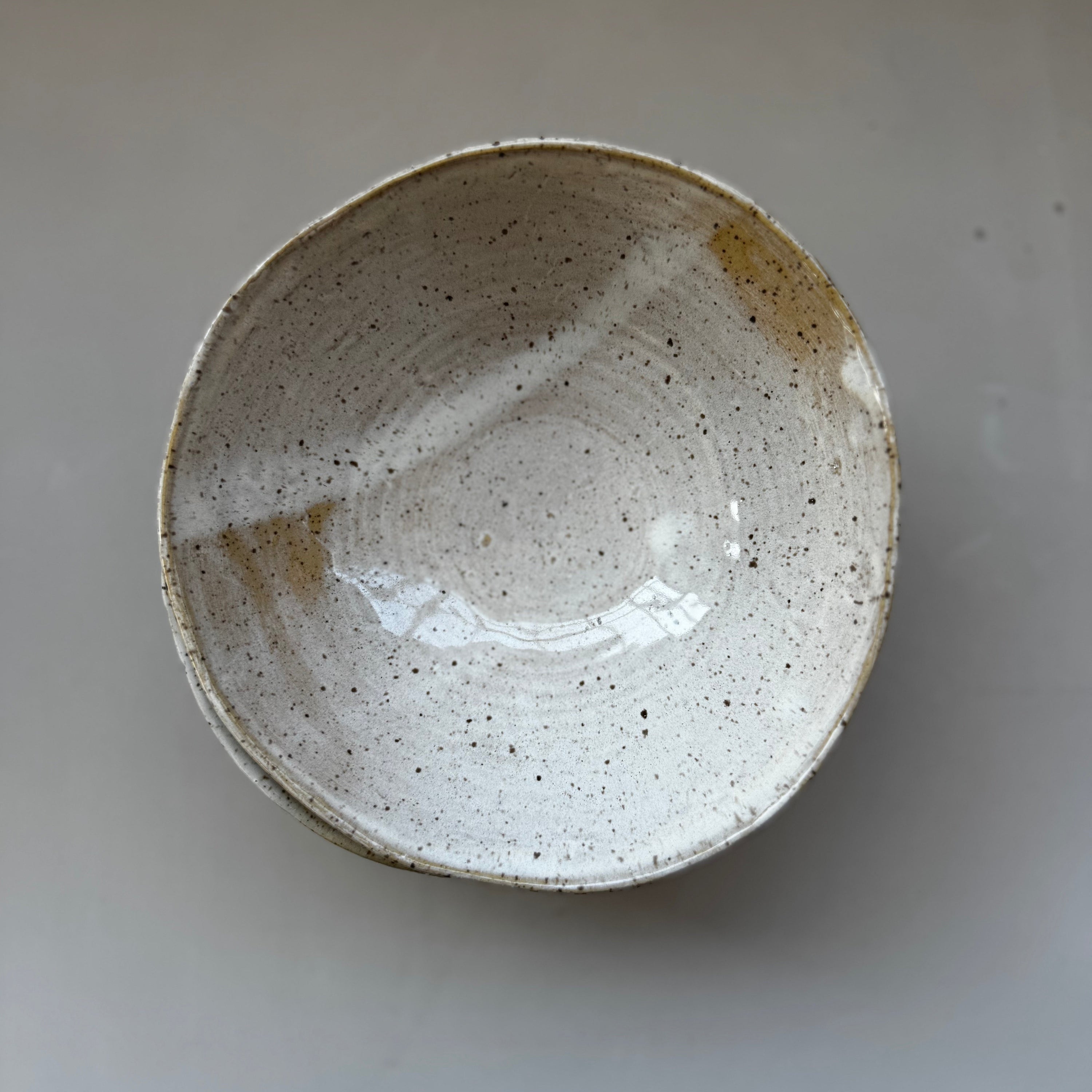 A handmade ceramic bowl with a speckled off-white glaze and a rustic brown rim, photographed from directly above on a neutral gray background.