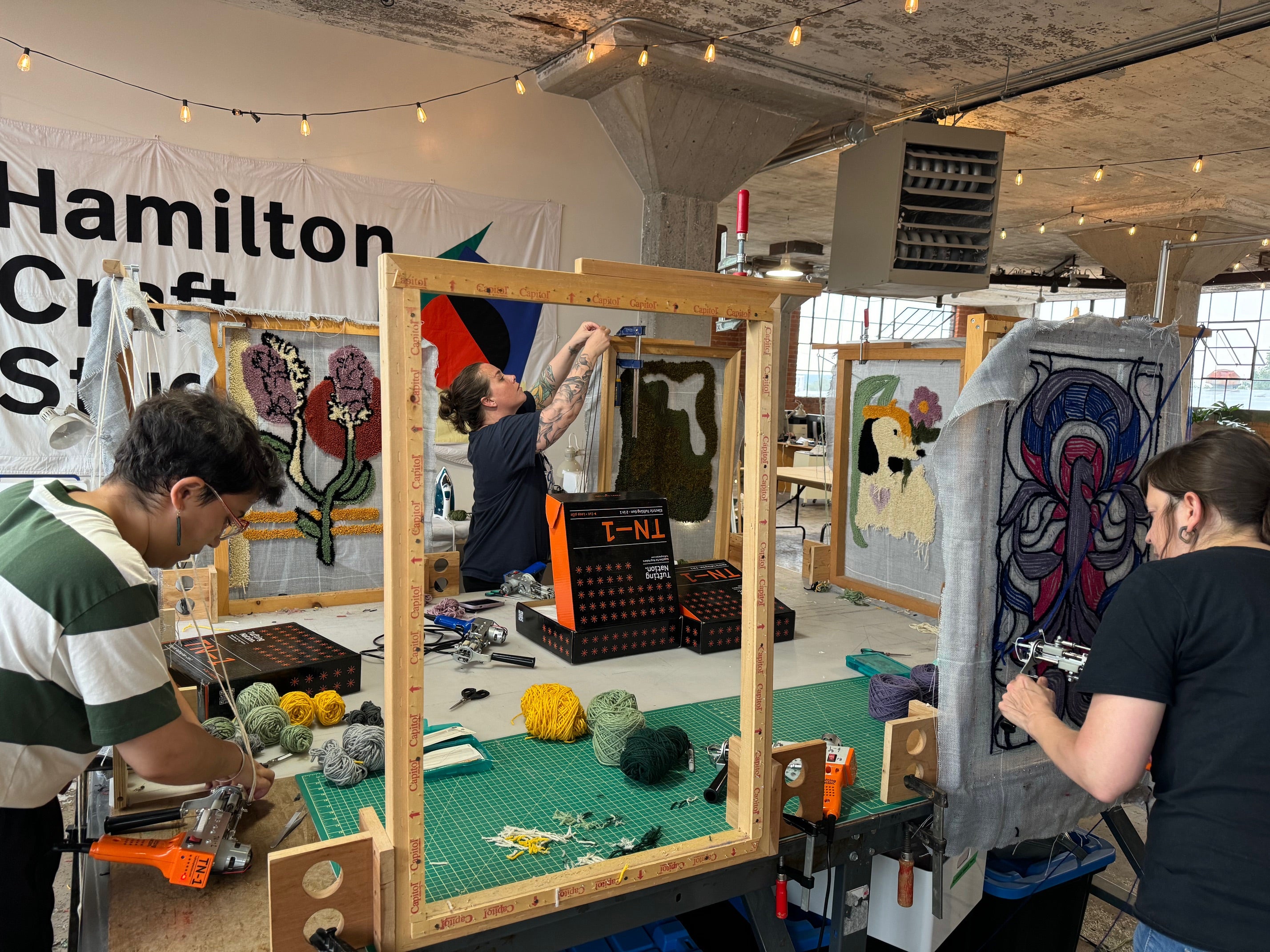 Art studio with people working on rug tufting projects, including an empty rug tufting frame and colorful rug designs on surrounding frames.