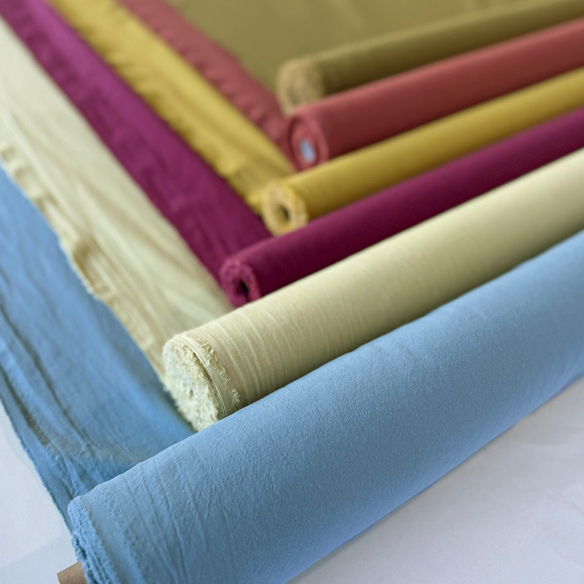 Multiple rolls of fabric in assorted colors, including light blue, pale yellow, magenta, mustard yellow, and olive green, arranged diagonally on a white surface.