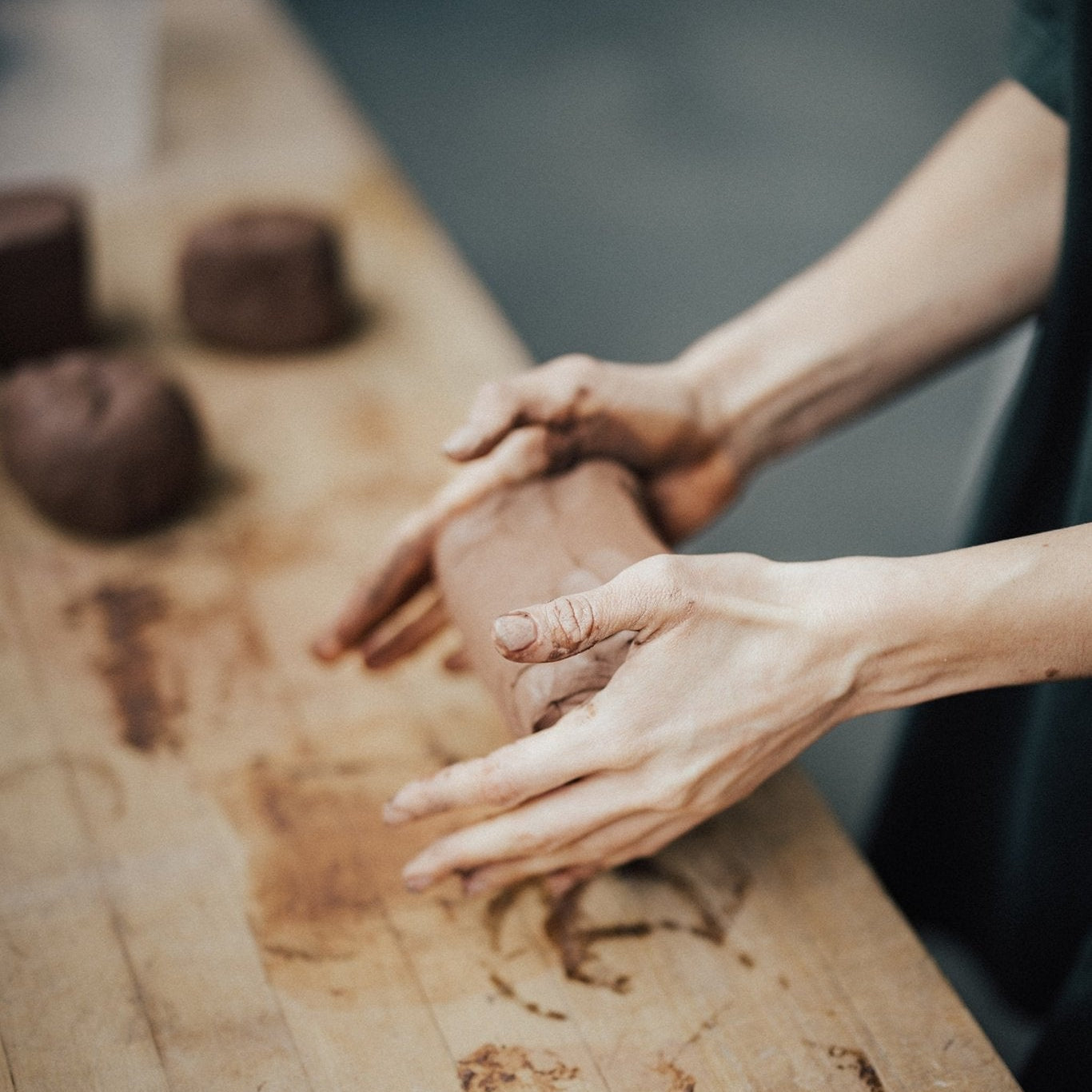a woman's hands wedging and preparing rad clay for throwing, on a wood surface with clay stains and wedged clay ready