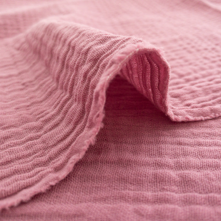 A close-up of a dusty pink textured muslin fabric, folded over to show its ribbed weave and soft, slightly sheer quality.